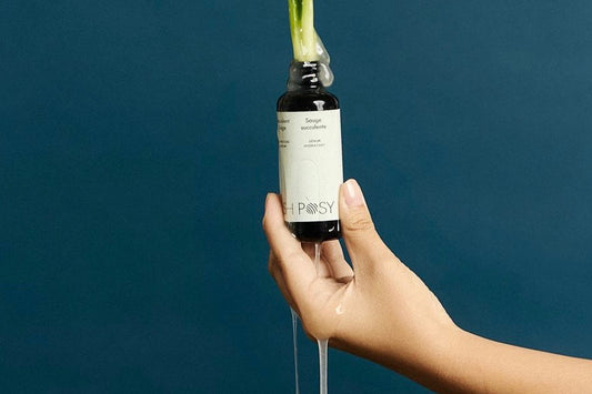 Hand holding a dropper bottle labeled "ASH POSY" against a blue background