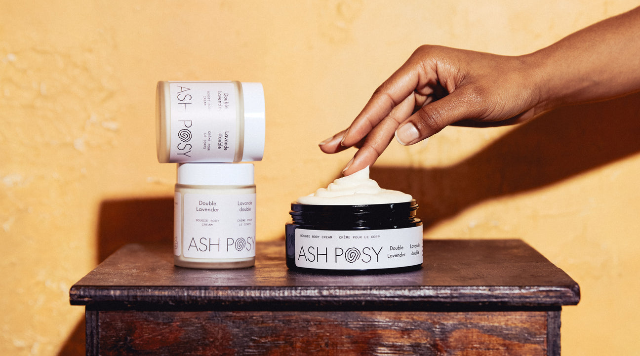 Hand applying cream from an 'ASH POSY' jar on a wooden surface with a yellow background